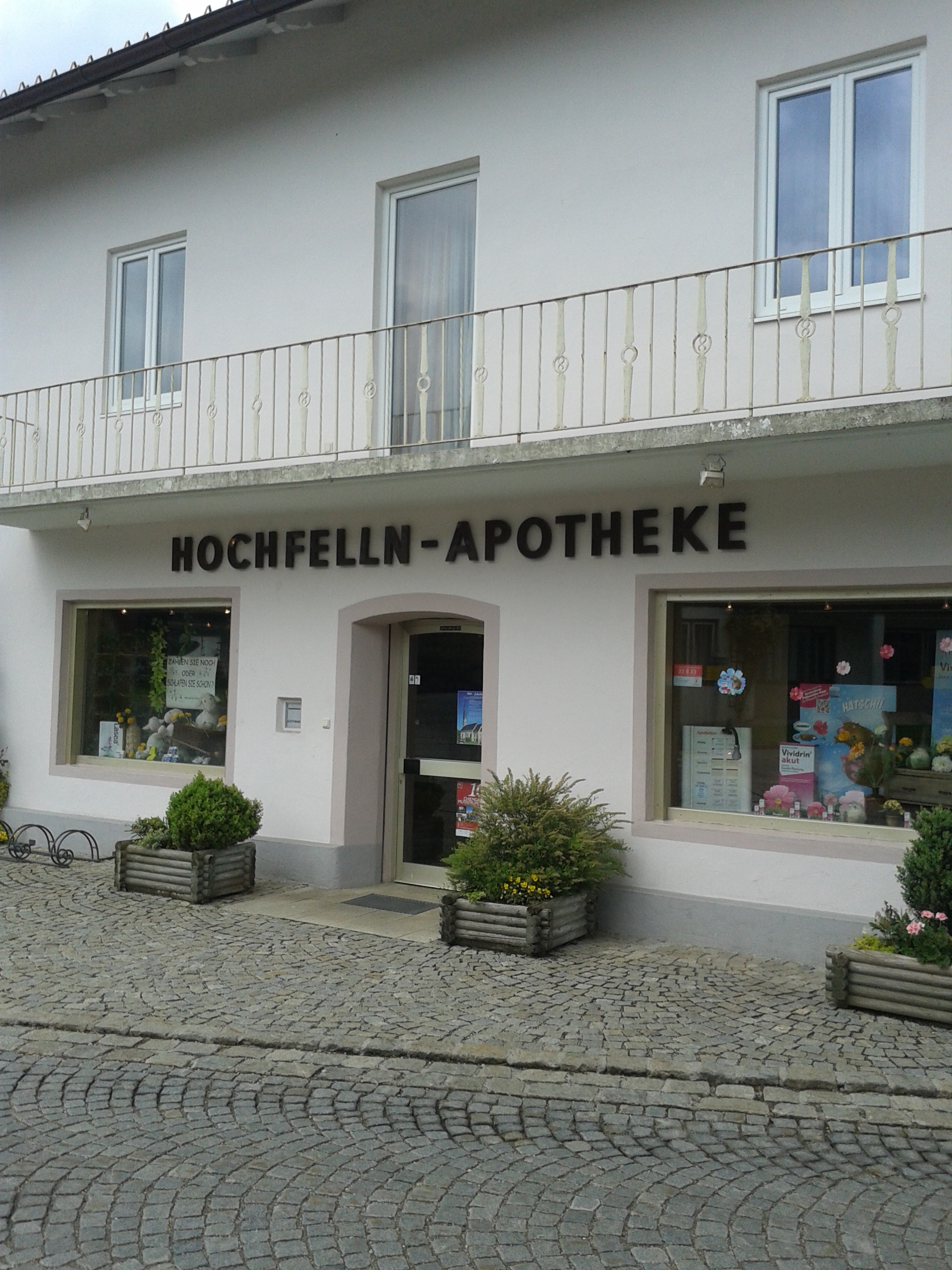 Zu Sehen ist die Seitenansicht eines hellen Hauses,in dem sich die Apotheke befindet. Vor dem Haus Blumenkübel und Fahrradständer,in den Schaufenstern die Auslagen mit Angeboten. Vor der Apotheke ist die Dorfstrasse zu erkennen.