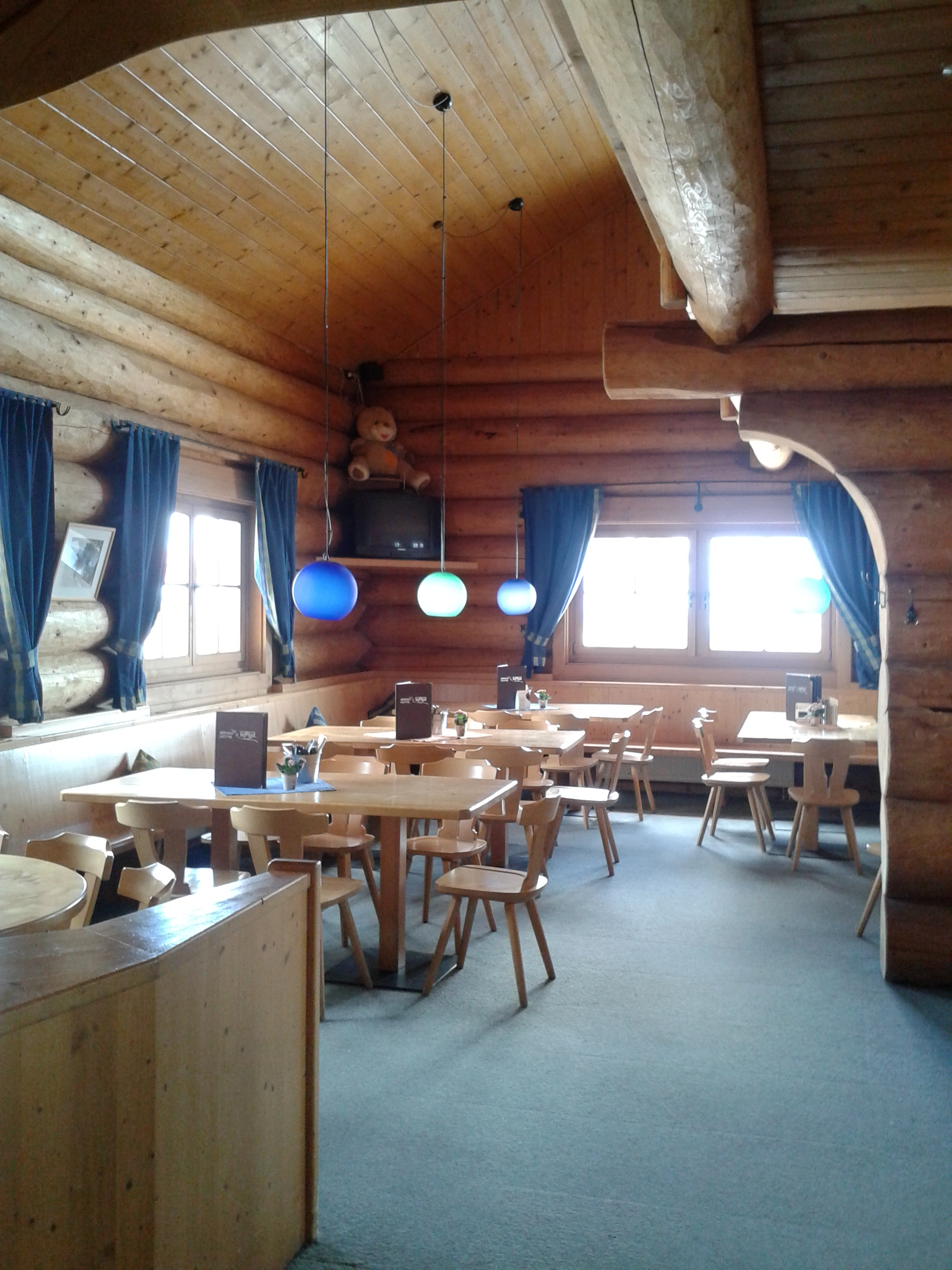 Das Innere einer Holzblockhütte, Tische und Stühle sind aus hellem Holz, die Schänke ist mit hellblauem Teppichboden ausgelegt, vor den kleinen Fenstern befinden sich dunkelblaue Vorhänge. Auf den Tischen stehen Speisekarten, in einer Ecke des Raumes hängt ein Fernseher.