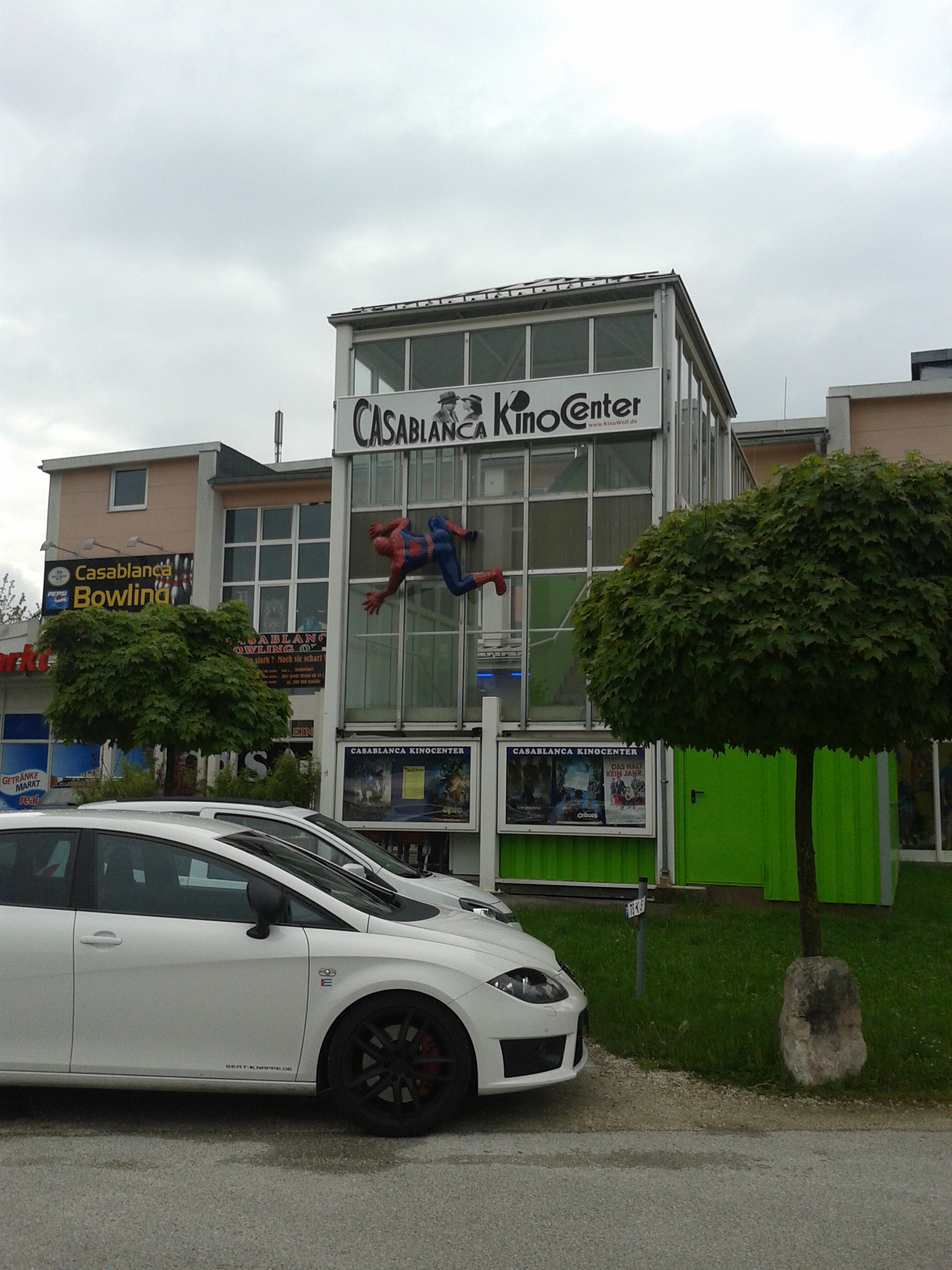 An der Dorfstraße stehen geparkte Autos, hinter den Autos ist ein modernes, verglastes Kinogebäude mit Plakatwänden, auf denen sich Filmhinweise befinden. An der Glasfassade ist die Figur von Spiderman angebracht. Links und rechts zeigen sich verschiedene Geschäfte und eine Bowlingbahn.